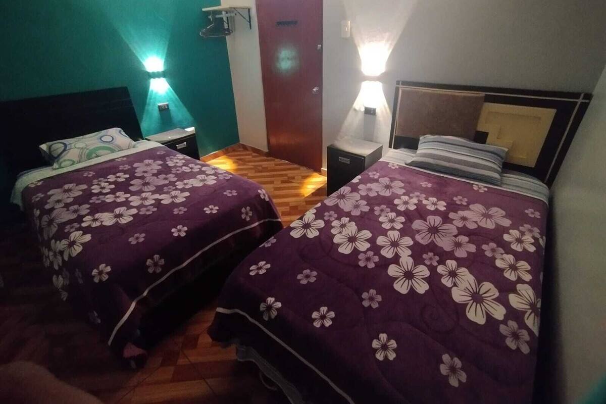A room with two beds, each with a purple floral bedspread, a wooden floor, a teal wall, and a wooden door.