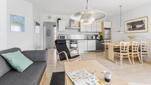 A modern apartment with a grey sofa, a dining table with chairs, a kitchen with white cabinets, and a large pendant light. - Gudhjem Søpark Lejl. 42 (Gudhjem)