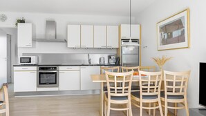 A modern kitchen with white cabinets, a microwave, a stove, and a dining area with a table and chairs. - Gudhjem Søpark Lejl. 42 (Gudhjem)