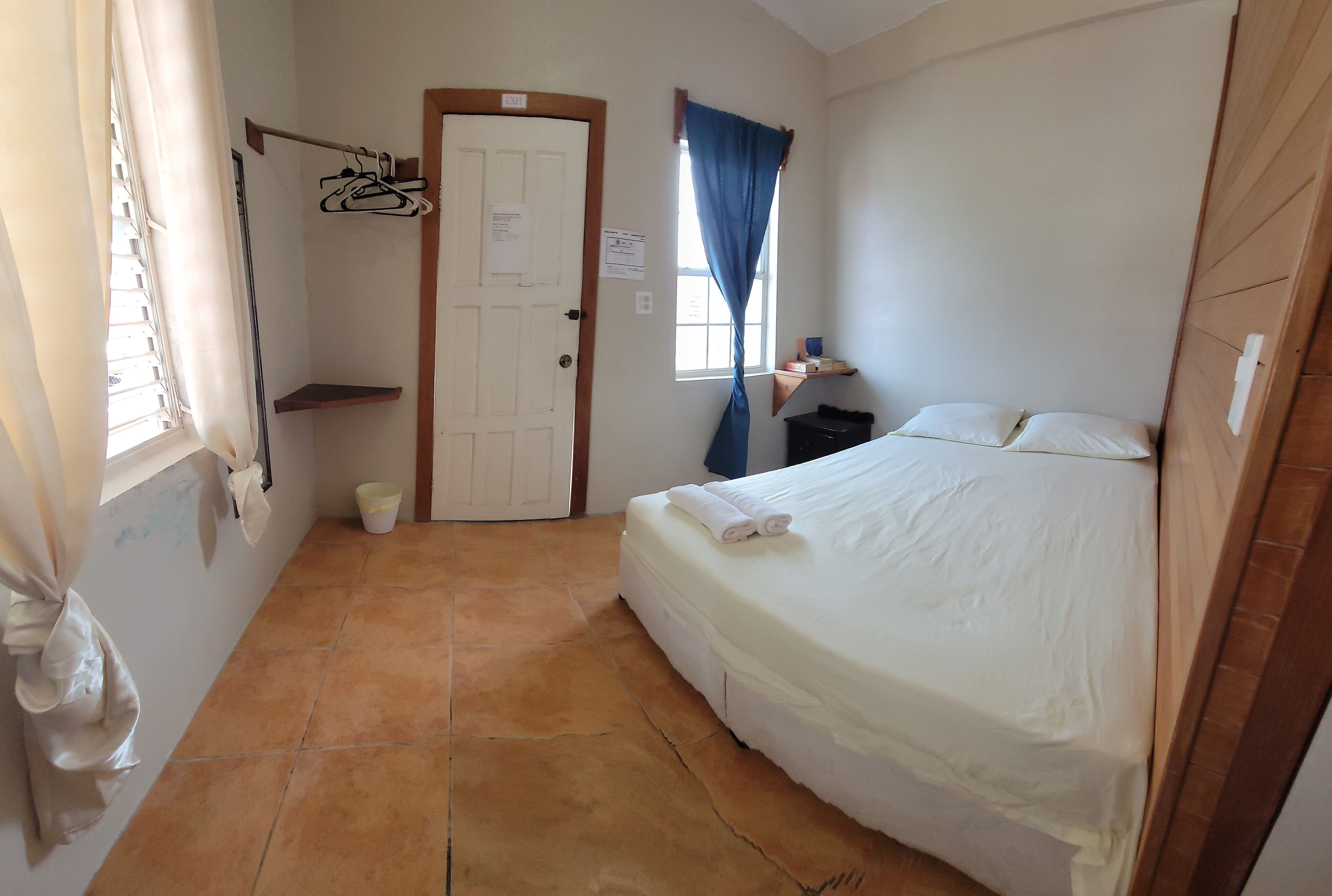 Una habitación pequeña y limpia con una cama, una puerta, una ventana con cortinas y un estante pequeño.