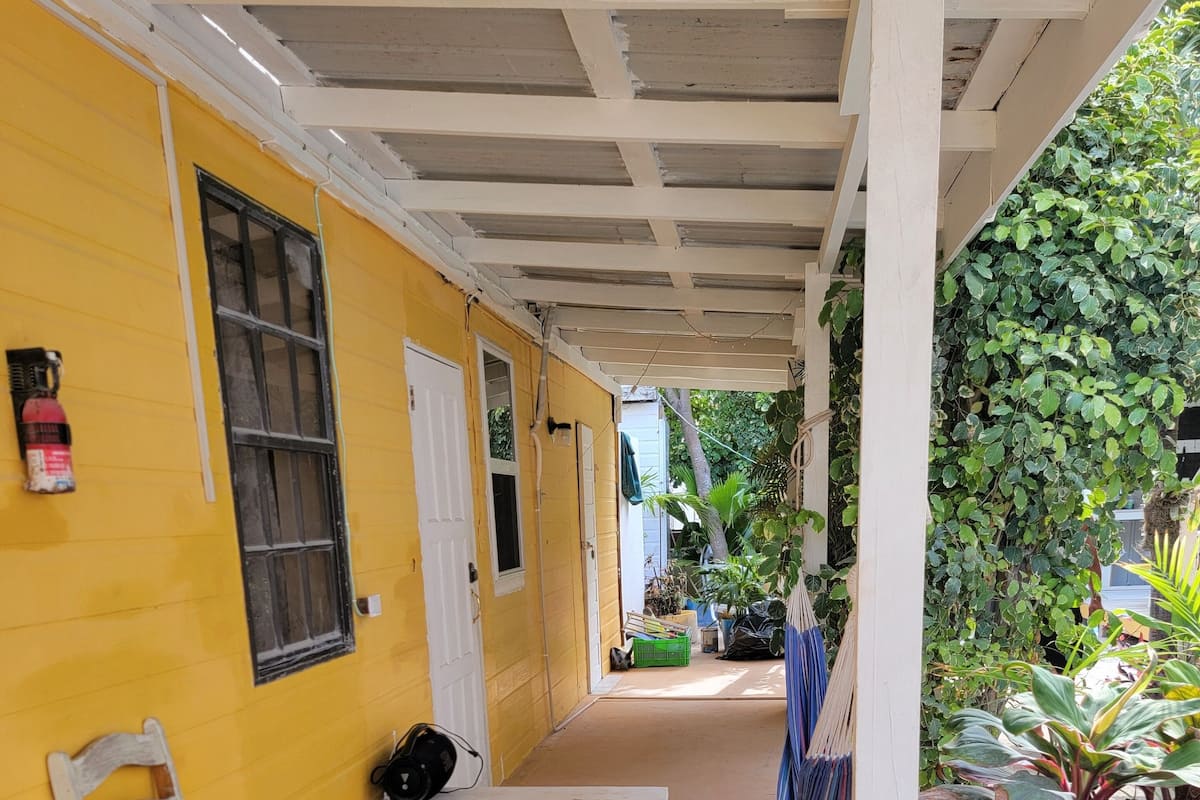 A covered outdoor area with a yellow wall, a wooden bench, a chair, and a fire extinguisher.