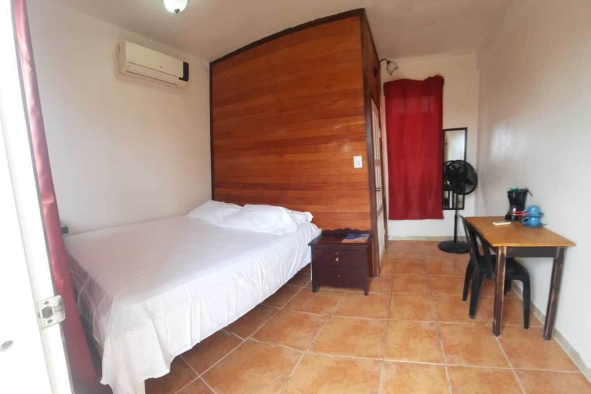 A bedroom with a wooden headboard, a bed, a small table with a fan, and a red curtain.
