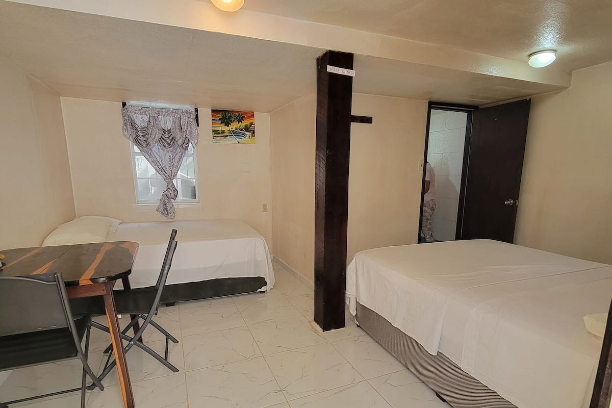 A small, clean room with a bed, a desk, and a chair. There is a window with a view of palm trees and a painting on the wall.