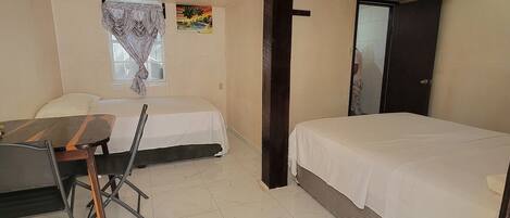 A small, clean room with a bed, a desk, and a chair. There is a window with a view of palm trees and a painting on the wall.