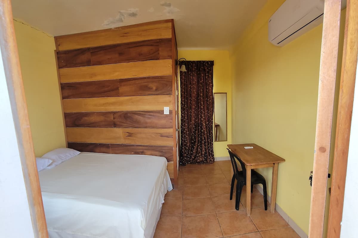 A room with a wooden headboard bed, a small table with a chair, and an air conditioning unit on the wall.