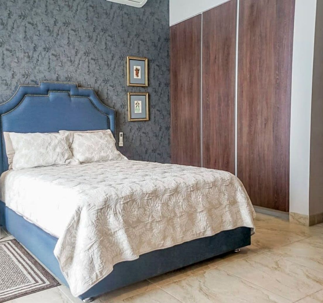 Un dormitori amb un llit, un capçal blau fosc, una paret amb textura gris i un armari de fusta.