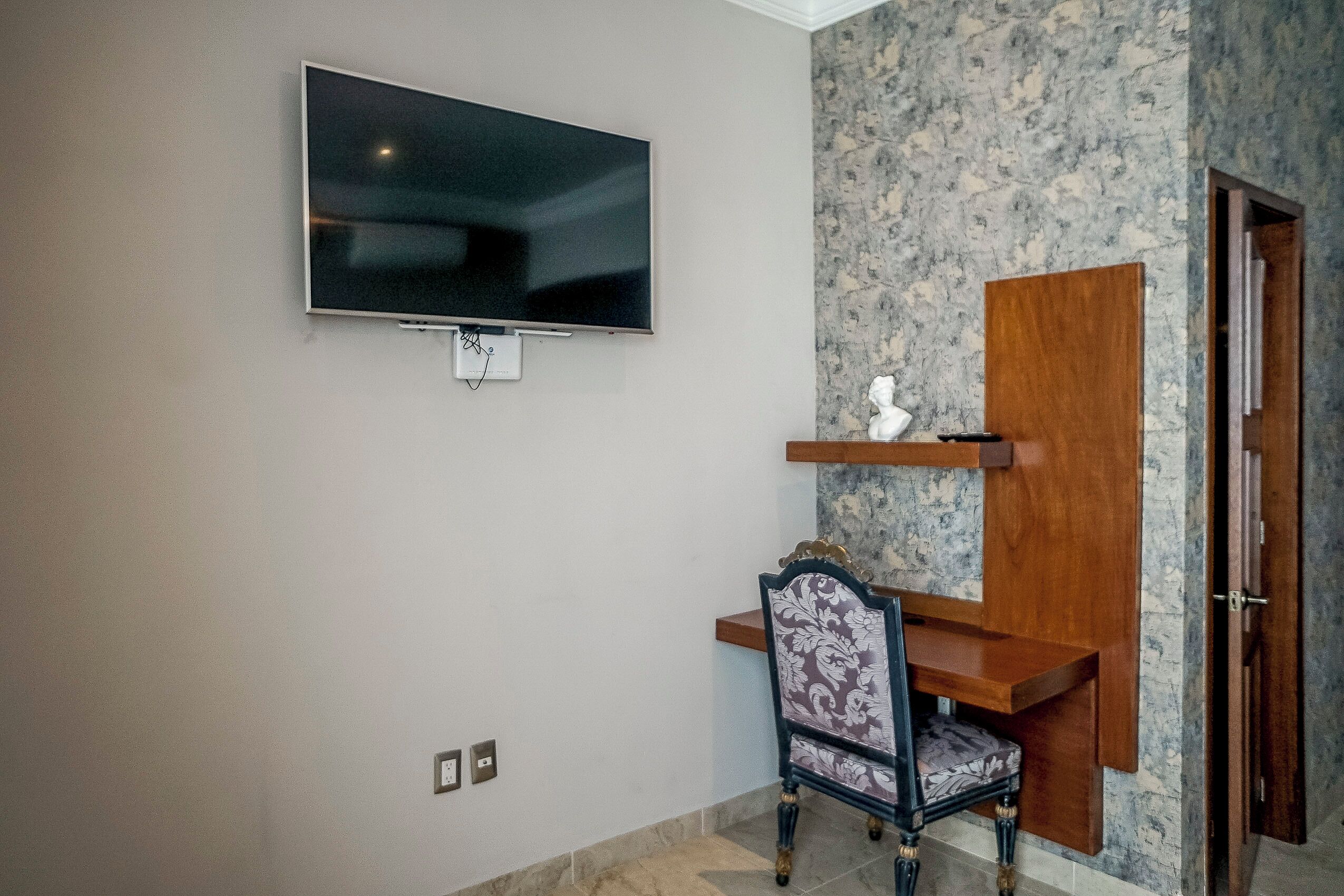 A room with a flat-screen TV mounted on the wall, a wooden desk with a chair, and a marble-patterned wall.