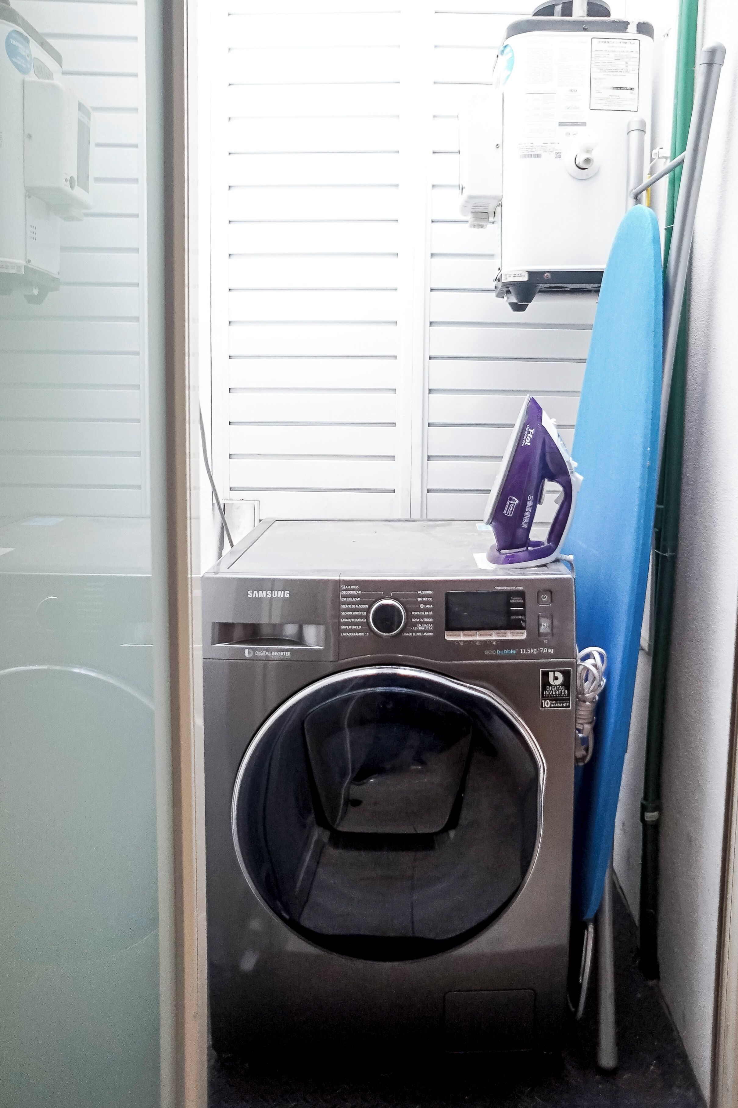 A compact, stainless steel Samsung digital washer and dryer unit in a small laundry room with a blue ironing board leaning against the wall.