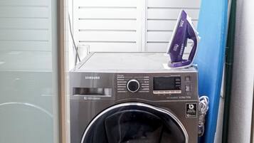 A compact, stainless steel Samsung digital washer and dryer unit in a small laundry room with a blue ironing board leaning against the wall.