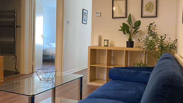 A modern living room with a blue sofa, a glass coffee table, and a wooden side table. There are two framed pictures on the wall and a potted plant on the side table. - Cosy 2 Bedroom Gem near Oxford Street (London)