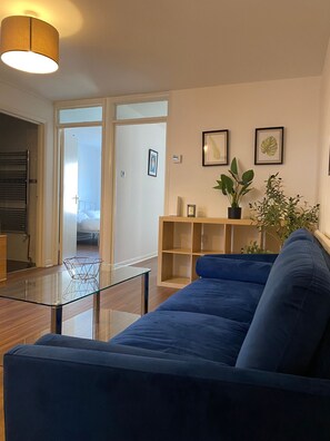Una sala de estar moderna con un sofá azul, una mesa de centro de vidrio y una mesita de noche de madera. Hay dos cuadros en la pared y una planta en maceta sobre la mesita.