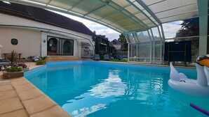 An indoor swimming pool with a glass enclosure, a swan float, and a patio area with seating and plants.