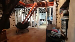 A spacious room with a wooden floor, a black chair, a staircase with a red railing, and a white wall with a patterned design.