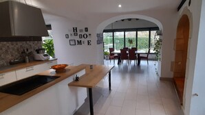 A modern kitchen with a central island, a dining area with a table and chairs, and a view of greenery through large windows.