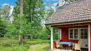 A red wooden house with a tiled roof, a porch with wicker furniture, and a view of a lush green forest. - Nice home in Annerstad with WiFi (Annerstad)