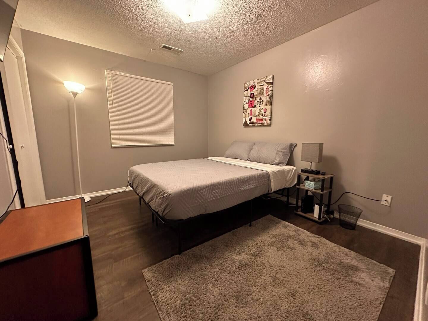 Une chambre à coucher comprenant un lit, une table de chevet, une lampe, un tableau décoratif et une fenêtre avec des stores.