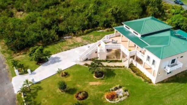 Un edificio de dos pisos con techo verde, un corredor cubierto y un jardín ajardinado con palmeras.