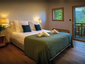 A neatly made bed with a green bedspread, two white pillows, and two dark green pillows. There are two bedside lamps with white shades, a small wooden nightstand with a plant, and a view of a balcony with a wooden railing. - Chalet Lezopau - mountain break with spa and petanque - OVO Network (Samoëns)