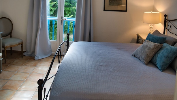 A bedroom with a bed, a nightstand, a lamp, a chair, and a window with curtains. - Orient Parc - the best location (Saint martin)