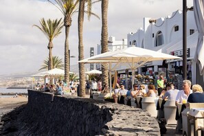 A coastal area with outdoor seating, palm trees, and a view of the beach and cityscape. - Home2Book Radiant Apt, Pool & Terrace Las Américas (Playa de la Américas)
