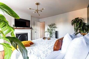 A modern bedroom with a bed, white bedding, a television above a fireplace, and potted plants. - Jungle House Urban Lofts – Arena District & Short North (Columbus)
