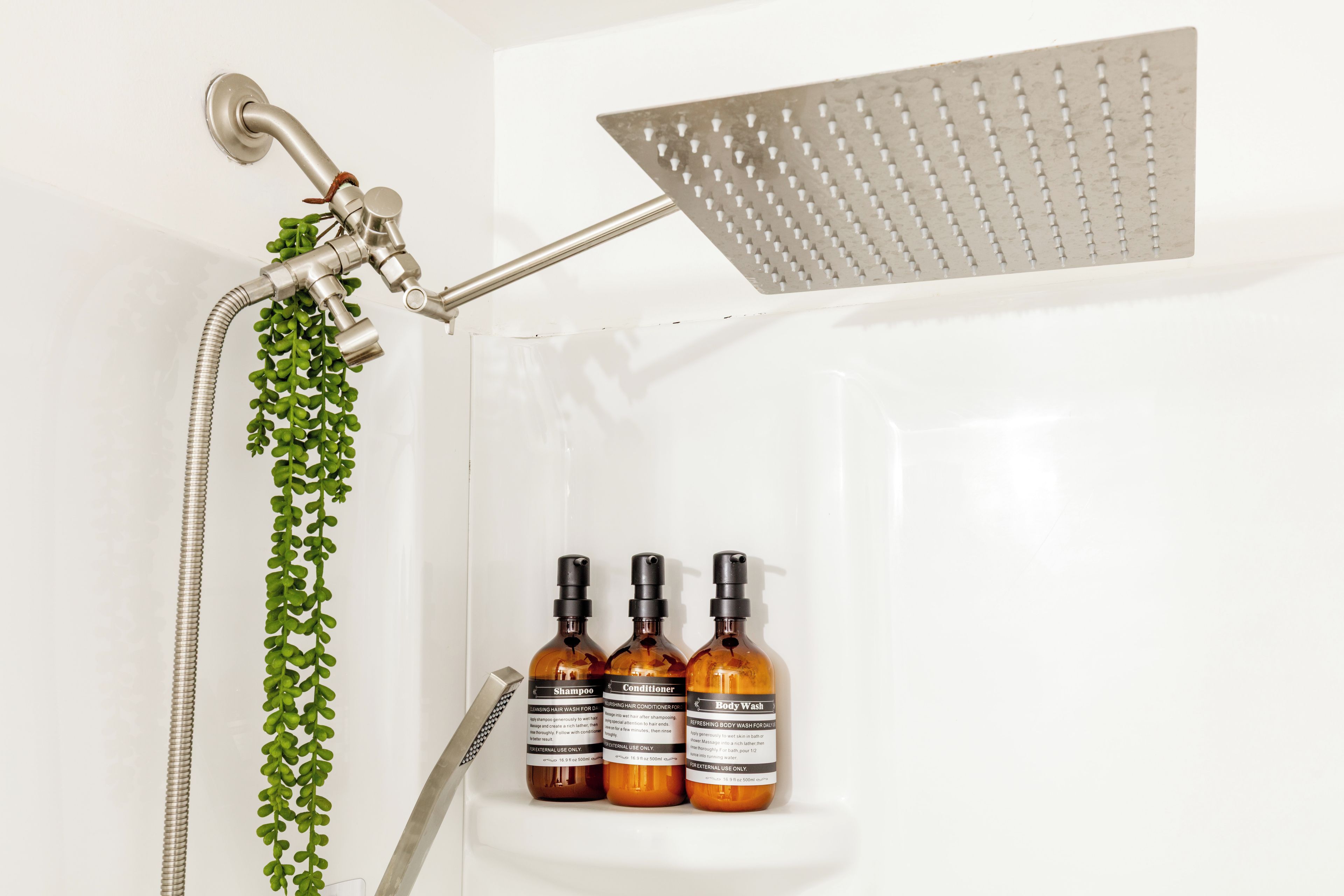 Ducha con cabezal de ducha de mano, cabezal de ducha de lluvia empotrado en la pared y una planta. Hay tres botellas de jabón corporal, champú y acondicionador sobre una repisa.