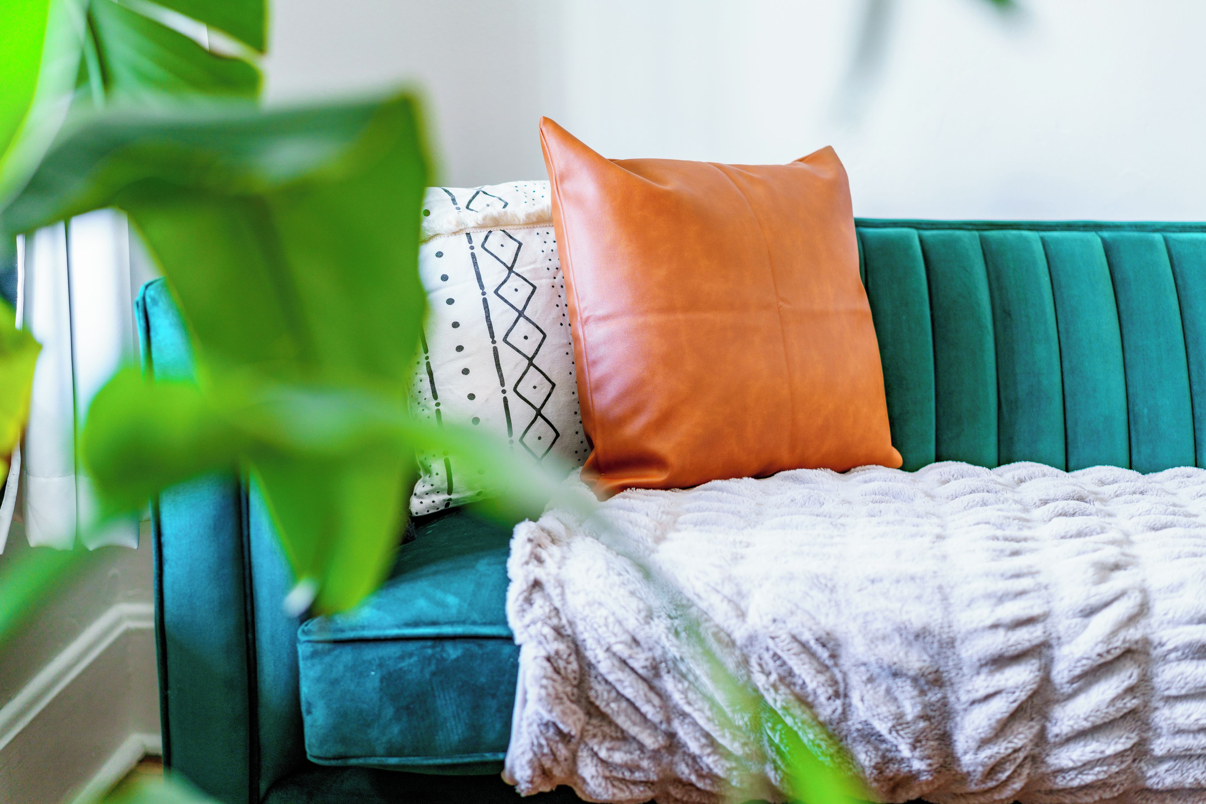A teal sofa with a patterned pillow and a solid orange pillow, a white knitted throw, and a green plant in the foreground.