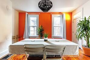 Un coin repas avec une table dressée pour deux personnes, deux fenêtres avec des rideaux, une plante en pot et un luminaire au plafond.