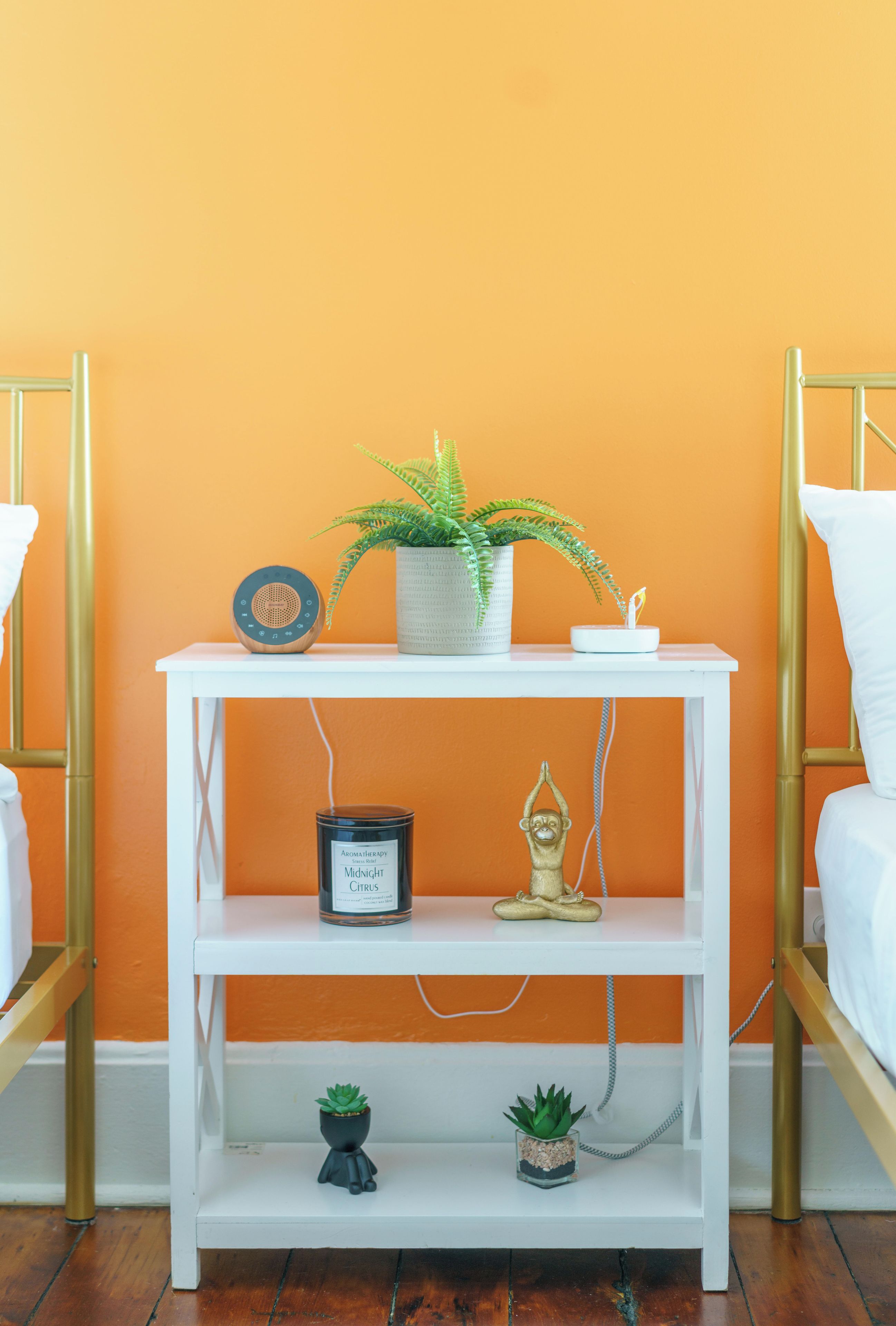 Ein Nachttisch in Weiß mit Blumentopf, Kerze und kleiner Figur vor einer orangefarbenen Wand.