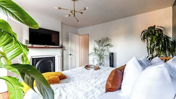 A modern bedroom with a bed, white bedding, a television above a fireplace, and potted plants.