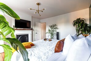 A modern bedroom with a bed, white bedding, a television above a fireplace, and potted plants. - Jungle House Urban Lofts – Arena District & Short North (Columbus)