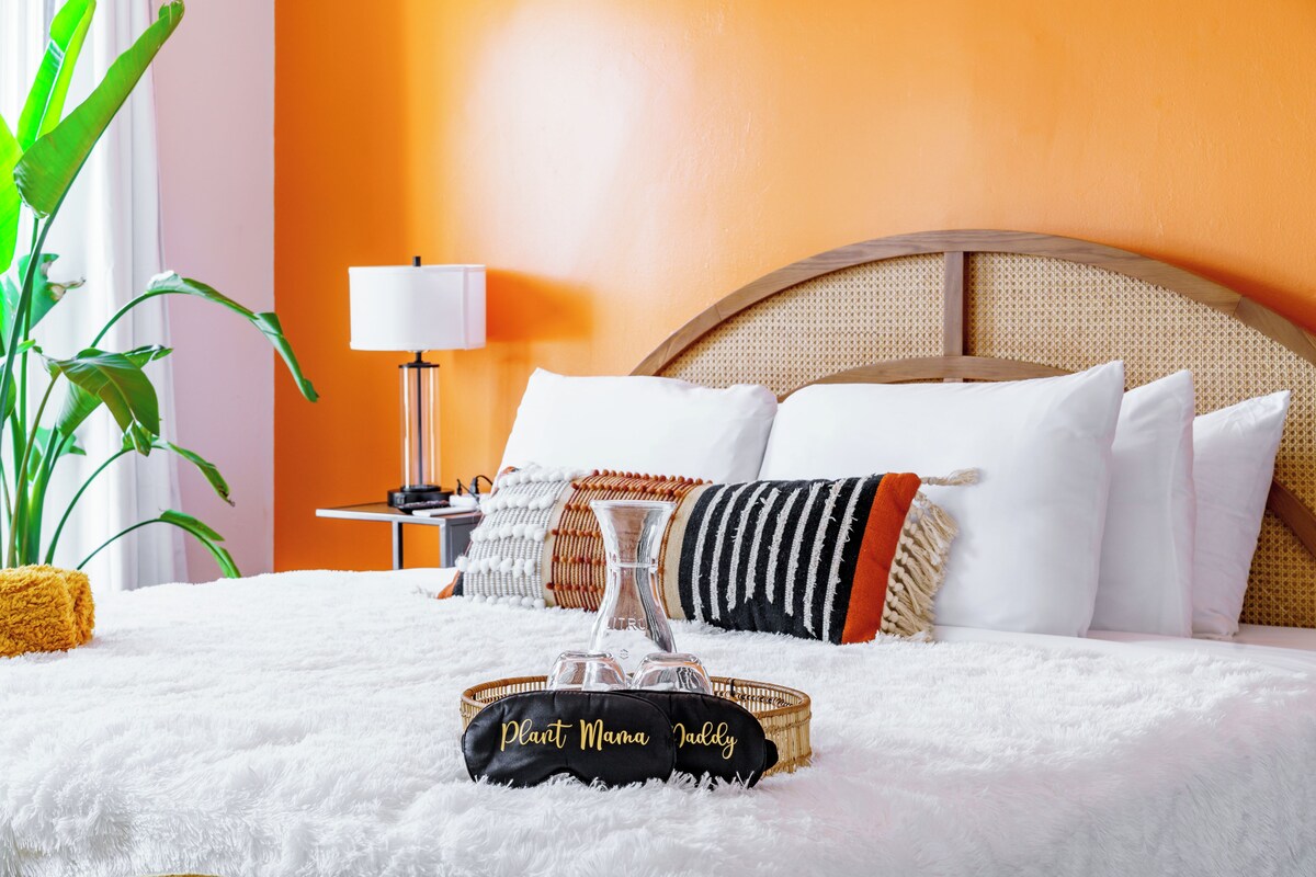 A hotel room with a bed, a bedside lamp, a potted plant, and a decorative tray with the text 'Plant Mama Jabbly'.