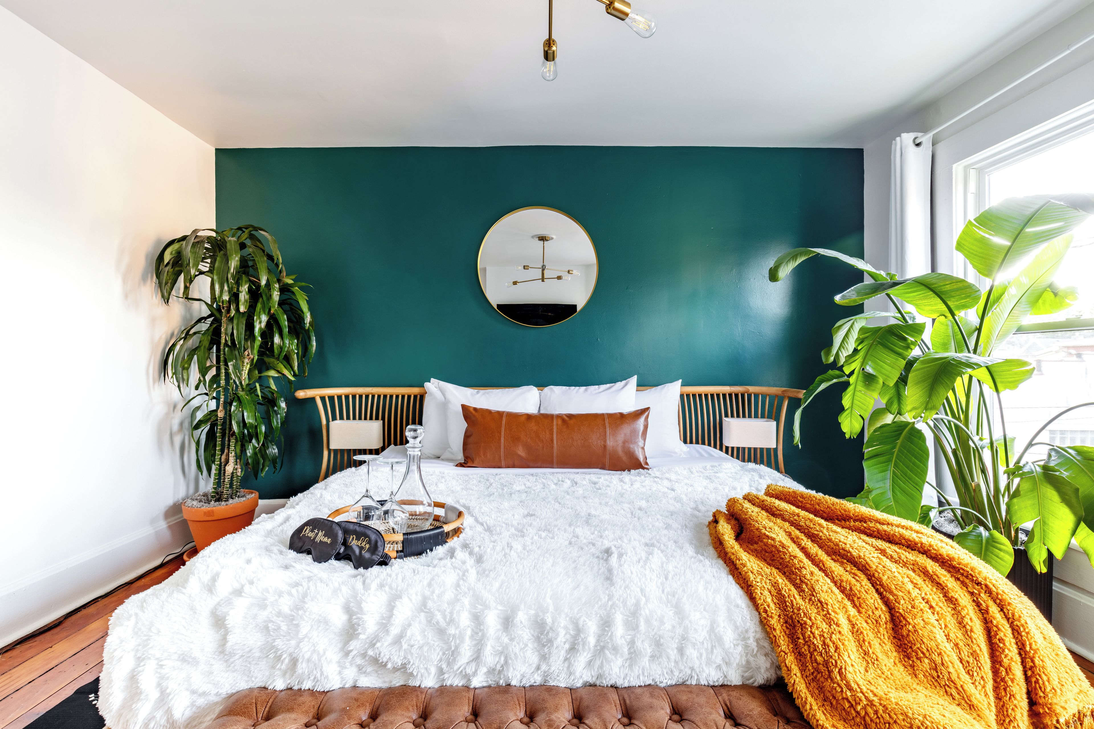A bedroom with a teal wall, a round mirror, a bed with white and brown bedding, and two potted plants.