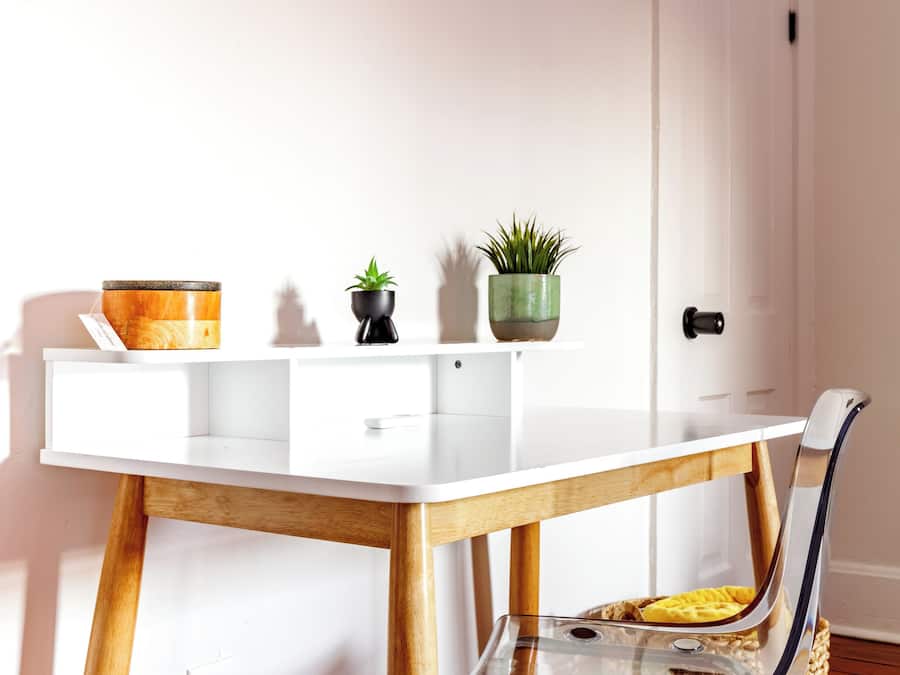 Een wit bureau met een glazen blad, houten poten en een stoel met metalen frame. Op het bureau staan twee planten in potten.