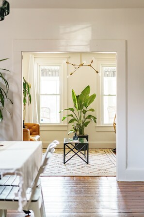 Ein Zimmer mit Esstisch, Stühlen, einer Topfpflanze, einer Pendelleuchte und einem fenster mit Vorhängen.