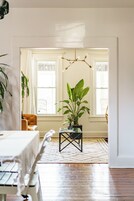 Una habitación con una mesa de comedor, sillas, una planta en maceta, una lámpara colgante y un ventanal con cortinas.