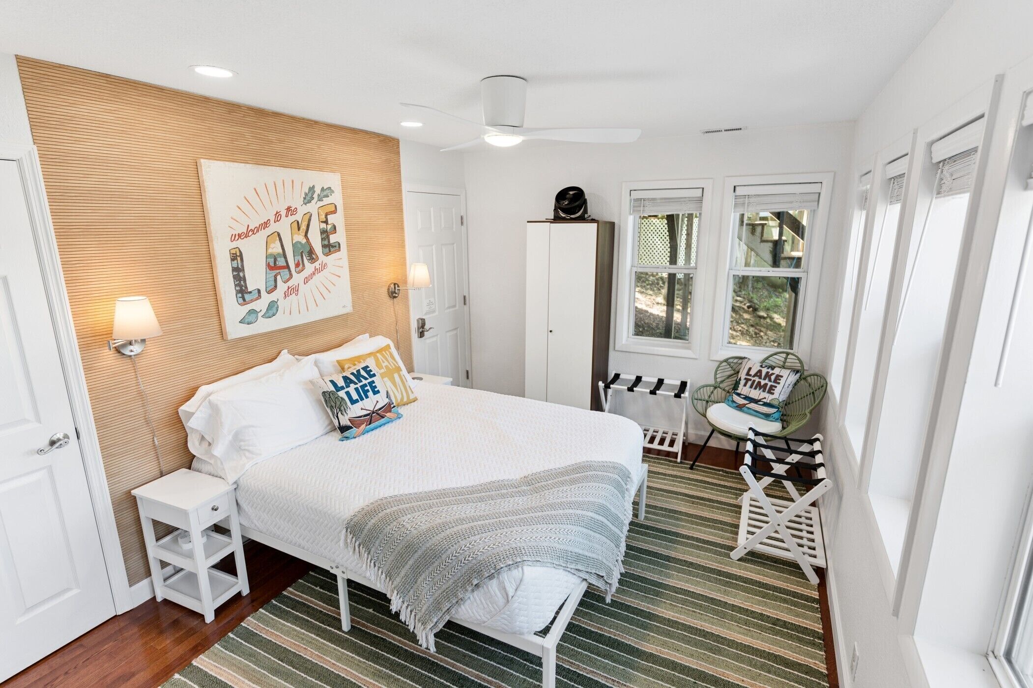 Un dormitorio con cama, mesita de noche, lámpara, decoración en la pared, armario, ventanas y una silla.