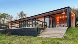 Een moderne houten hut met een grote glazen deur, buitenzitplaatsen en een vlag.