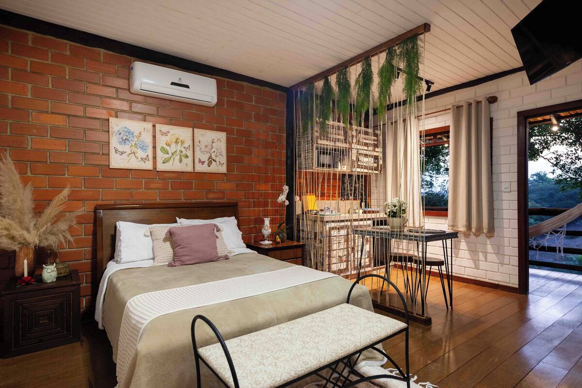 Une chambre avec un mur en briques, un lit avec une tête de lit, une table de chevet avec une lampe et une fenêtre avec des rideaux.