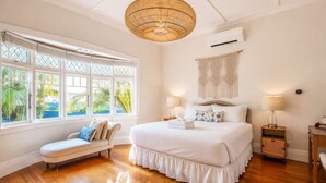 A bedroom with a large bed, a bench, a hanging light fixture, and a window with a view of greenery.