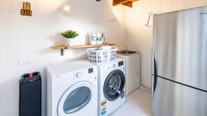 A laundry room with two washing machines, a refrigerator, a sink, and a shelf with a plant and laundry supplies.