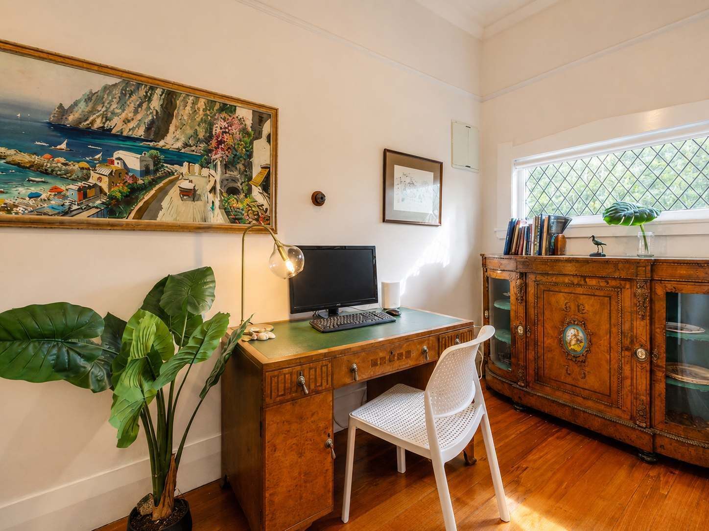 A room with a wooden desk, a computer, a white chair, a large potted plant, a wooden cabinet with glass doors, and a painting of a coastal scene.