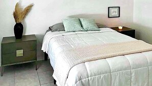 Une chambre à coucher avec un lit, une table de chevet, un vase contenant des plantes séchées et un tableau encadré accroché au mur.