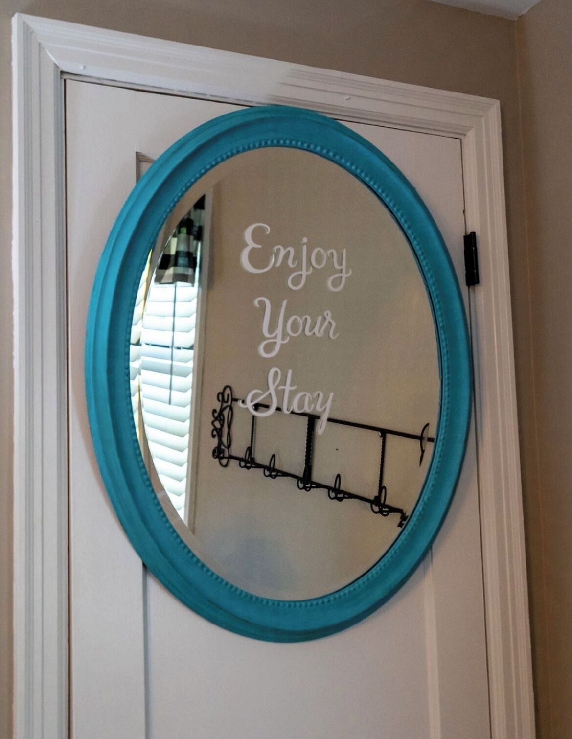 A mirror with a turquoise frame mounted on a white door, reflecting a window with blinds and a black coat rack with a sign that reads "Enjoy Your Stay."