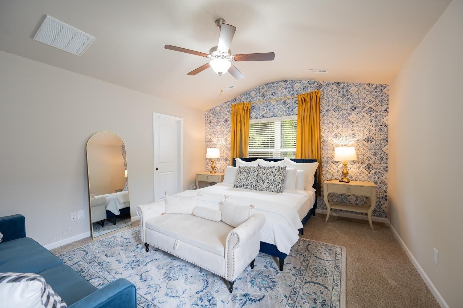 Une chambre à coucher avec un lit, un ventilateur de plafond, une table de chevet avec une lampe, un miroir et une chaise bleue.