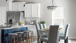 Una cocina moderna con armarios blancos, salpicadero azul y un comedor con sillas a rayas y una araña.