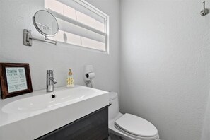A compact bathroom with a sink, mirror, and toilet, featuring a framed sign and amenities.