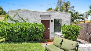 A small, single-story building with a white exterior and a brown door, surrounded by lush green bushes and a wooden fence. - 1BR/1BA Design District Cottage - 8 Min drive from Downtown WPB (West Palm Beach)