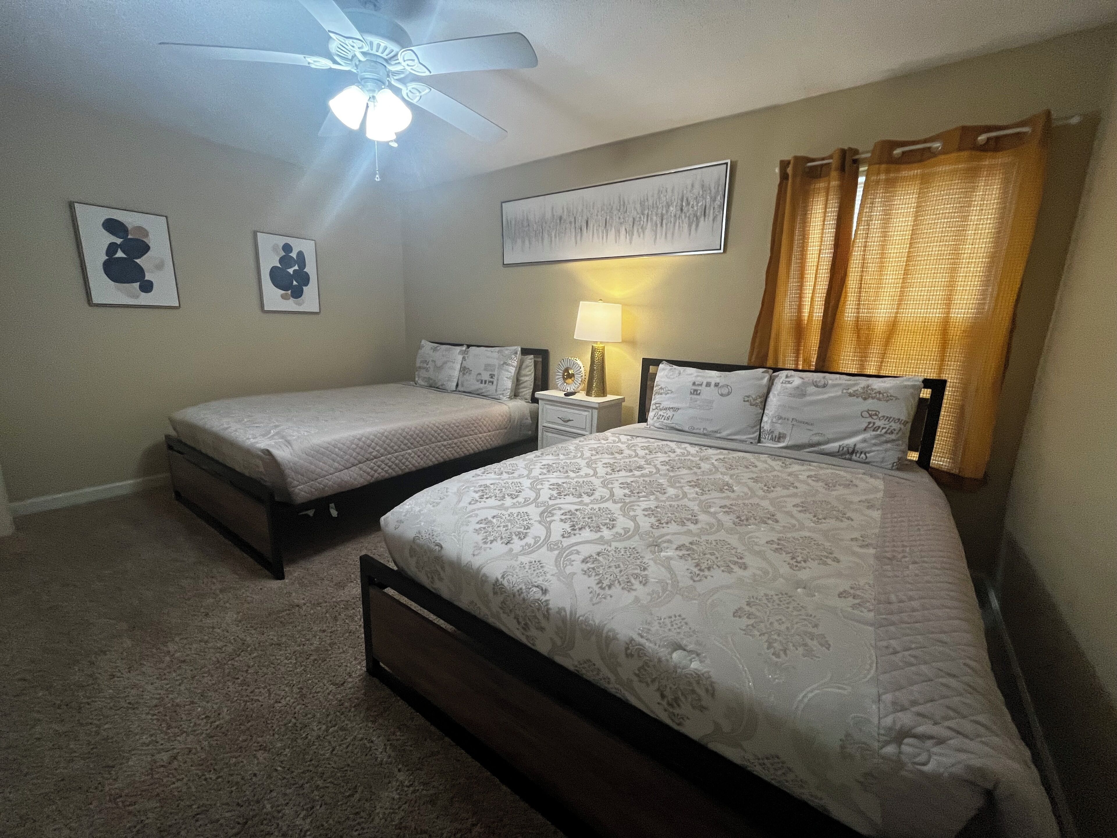 Une chambre avec deux lits, un ventilateur de plafond, une table de chevet avec une lampe et des œuvres d’art accrochées aux murs.
