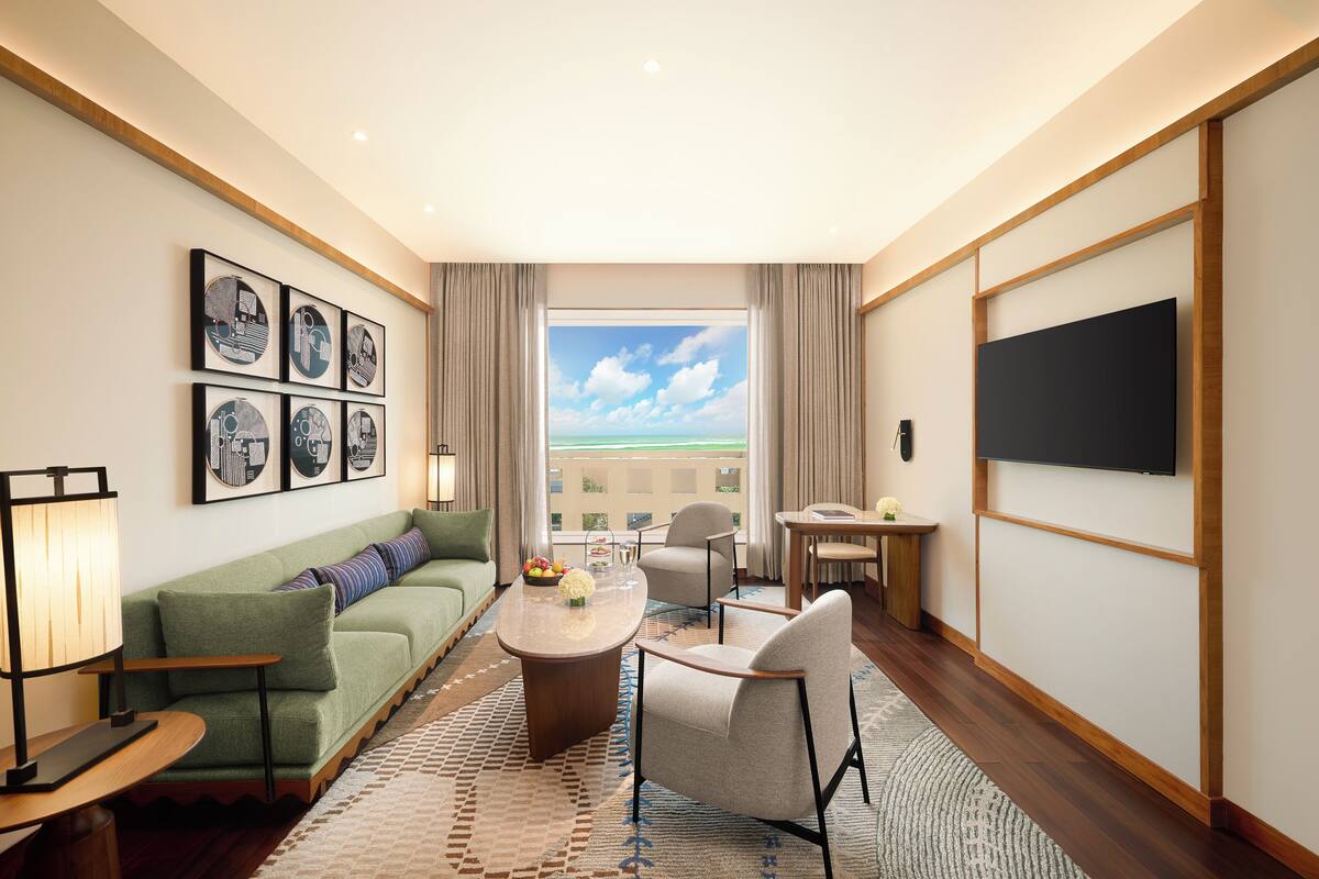 Hotelkamer met een groene bank, een houten salontafel, een televisie en een groot raam met uitzicht op de lucht en gebouwen.